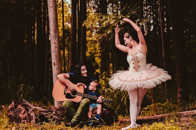 Un hombre en el bosque toca la guitarra mientras sostiene a un niño, junto a una bailarina bailando