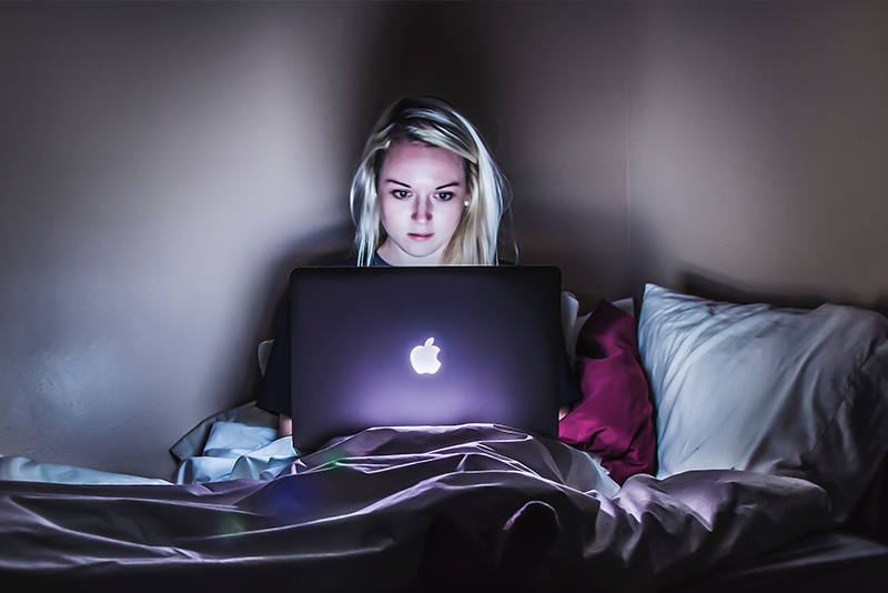 ผู้หญิงนอนอยู่บนเตียงมองไปที่หน้าจอแล็ปท็อปที่ส่องแสง
