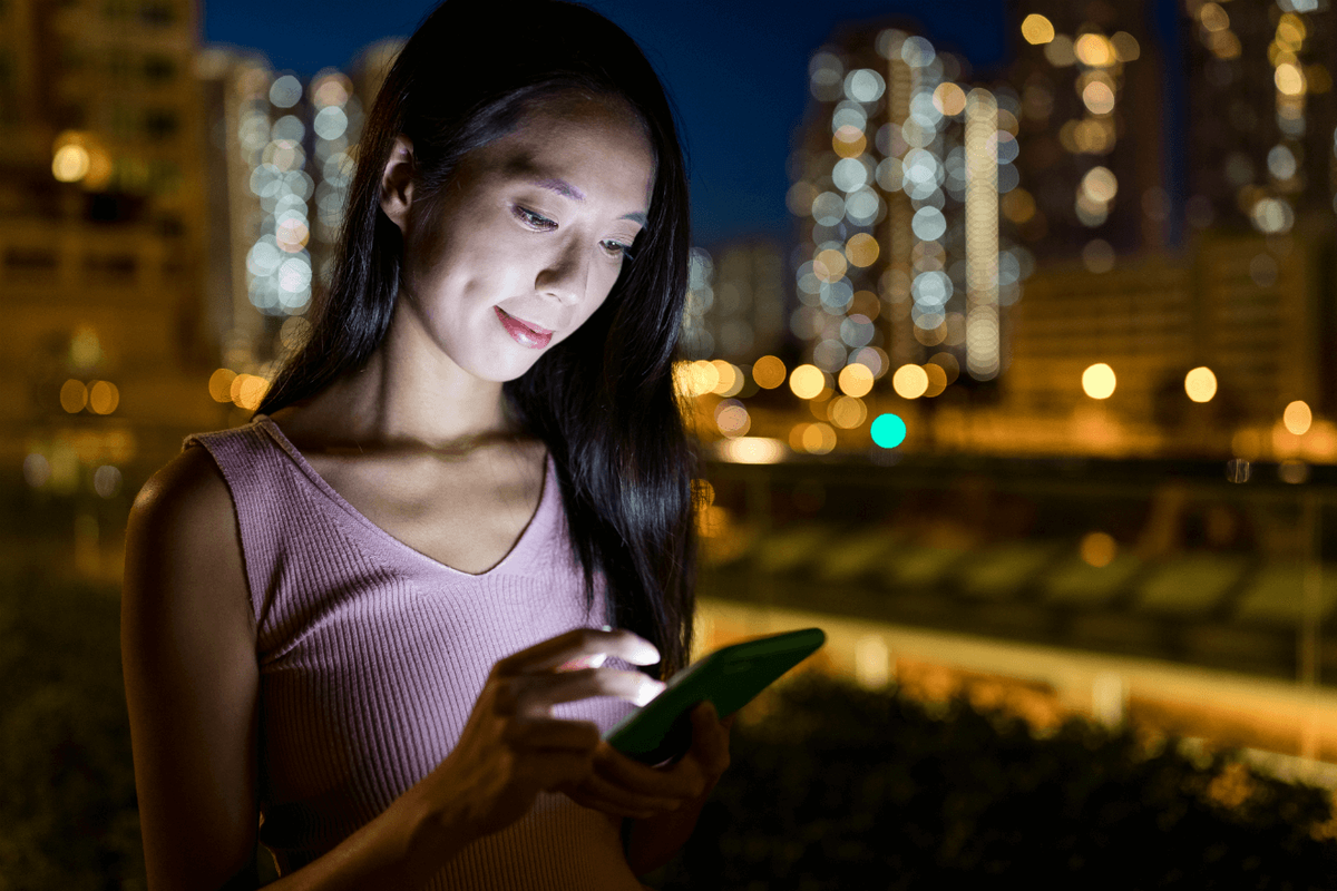 nainen tarkastelee älypuhelinta yöllä kaupungin taustalla